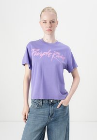 Purple cotton t-shirt featuring a pink graphic print reading "Purple Rain," with a relaxed fit and short sleeves. Paired with blue jeans.