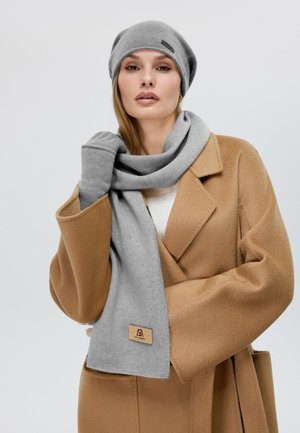 Un berretto in maglia grigia e una sciarpa abbinati a un cappotto di lana beige. La sciarpa presenta un'etichetta del marchio discreta; entrambi hanno una consistenza morbida e un design semplice.