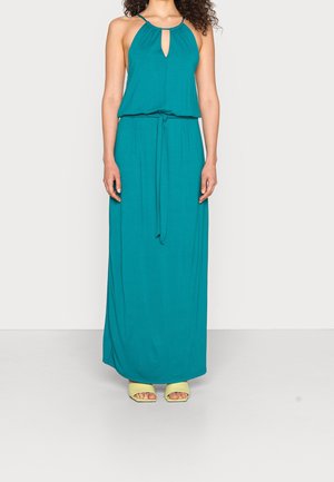 Vrouw draagt een teal halternek maxi-jurk met een tailleband, gecombineerd met lichtgroene open-teen sandalen met hakken, staand tegen een effen achtergrond.