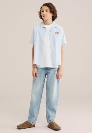 Bērns ar vidēja garuma brūniem matiem, valkā gaiši zilu svītrainu īsroku kreklu, baltu T-kreklu, vaļīgas, gaiši mazgātas džinsa bikses un brūnas ērti uzvelkamas kurpes.