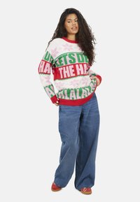 Maglione lavorato a maglia in rosa, rosso e verde con scritte festive e motivi a fiocchi di neve. Indossato con jeans a gamba larga e calzature rosse per un look casual.