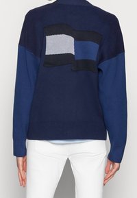 Suéter de punto azul marino con un diseño de bloques de color. Presenta un parche texturizado que se asemeja a una bandera en la parte posterior, con acentos en negro, azul y gris.