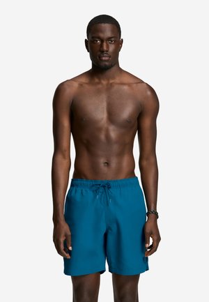 Bărbat în formă, stând drept, îmbrăcat în pantaloni scurți de baie albaștri cu șnur, pe un fundal simplu și luminos, fără tricou sau pantofi vizibili.
