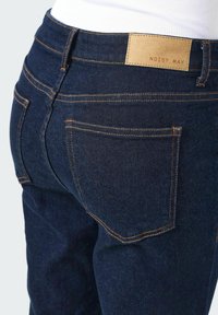 Temno modri jeans z oranžnim šivanjem, ki imajo posebne usnjene oznake z napisom "NOISY MAY" na pasu in zadnjih žepih.