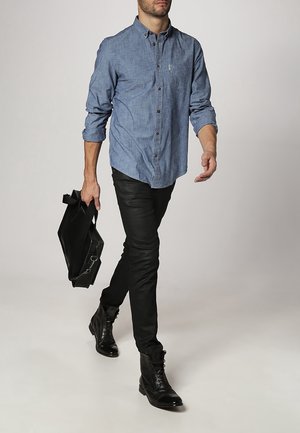 Lichtblauw denimoverhemd met knopen, lange mouwen opgerold, gecombineerd met zwarte spijkerbroek en zwarte veterboots. Draagt een zwarte tas.