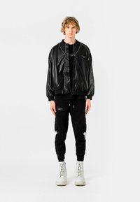 Chaqueta bomber de cuero negro con cremallera y bolsillos, combinada con joggers negros con detalles desgastados. Calzado son botas blancas con cordones.
