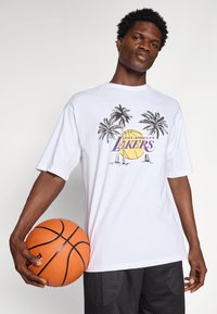 Biela tričko s krátkym rukávom, zdobené grafickým dizajnom s palmami, basketbalom a nápisom "Los Angeles Lakers" v purpurovej a žltej farbe.