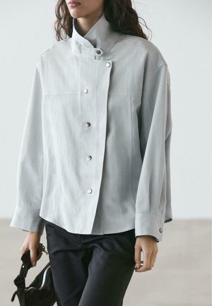 Giacca grigia chiaro con bottoni, collo alto, vestibilità ampia, maniche lunghe e bottoni a pressione argentati sulla parte anteriore e sui polsini.