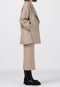 Beige oversized kappa i ullblandning med djupa slag, kombinerad med en ribbad beige midikjol. Svarta stövletter med tjock sula i ankellängd fulländar outfiten.