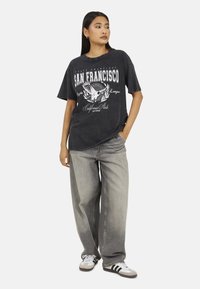 T-shirt grafica nera con stampa "San Francisco", maniche corte e vestibilità rilassata, abbinata a jeans grigi larghi e sneaker.