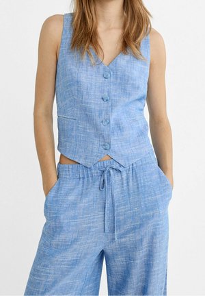 Woman wearing a sleeveless blue buttoned vest and matching drawstring wide-leg pants with hands in pockets.