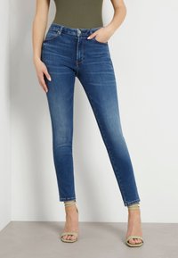 Jeans din denim albastru, mulate, cu talie medie, design cu cinci buzunare și o ușoară decolorare. Asortate cu sandale verzi cu barete.