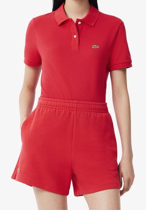 Kvinde iført rød polo t-shirt med korte ærmer og matchende røde elastiske shorts med krokodillelogo på begge beklædningsgenstande.