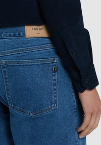 Jeans en denim d'une teinte bleu moyen avec une étiquette en cuir portant la mention "FARAH". Présente un design classique à cinq poches et des détails de couture subtils.