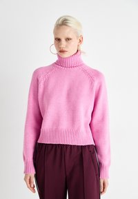 Pink knitted sweater with a high collar, ribbed cuffs and hem, featuring a loose fit and textured fabric. Paired with dark pants.