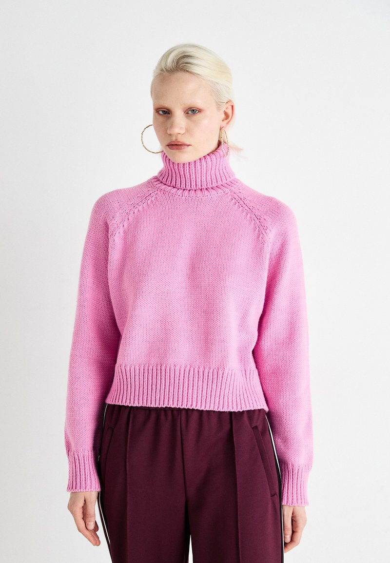 Pink knitted sweater with a high collar, ribbed cuffs and hem, featuring a loose fit and textured fabric. Paired with dark pants.