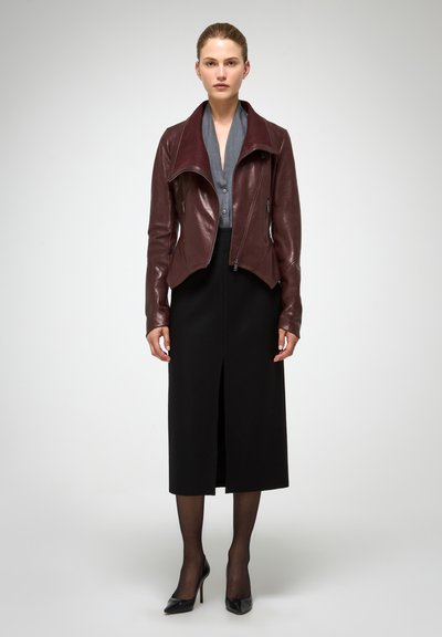 Veste en cuir bordeaux avec un large col, associée à un chemisier gris et une longue jupe noire, dotée d'une fente frontale et de talons noirs.