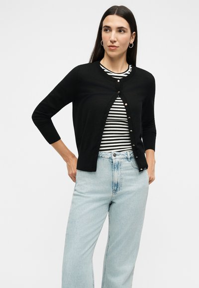 Γυναίκα με μακριά σκούρα μαλλιά που φοράει ασημένια σκουλαρίκια στεφάνες, μαύρη ζακέτα με κουμπιά πάνω από μαύρο-άσπρο ριγέ πουκάμισο και ανοιχτό μπλε τζιν.