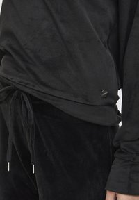 Maglione in pile nero con dettaglio regolabile all'orlo, caratterizzato da una texture liscia e un piccolo logo metallico accentato sul lato.