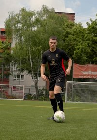 Svart idrottströja med röda detaljer, svarta shorts och knähöga strumpor. Spelaren står på en grön konstgräsplan och förbereder sig för att sparka en fotboll.