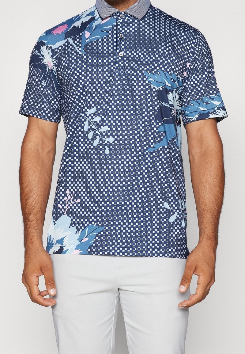 Polo de manga corta con base azul marino y patrones florales y geométricos contrastantes en azul y rosa, con un cuello texturizado.