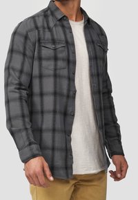 Chemise en flanelle à carreaux gris avec manches longues, fermeture boutonnée sur le devant, deux poches poitrine et un tissu texturé, portée sur un t-shirt de couleur claire.