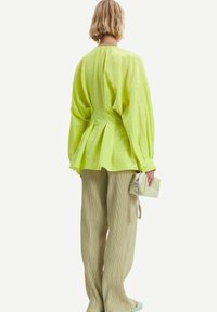Camicia oversize di un verde lime con texture e pieghe sulla schiena, abbinata a pantaloni beige chiaro a pieghe e una piccola borsa verde chiaro.