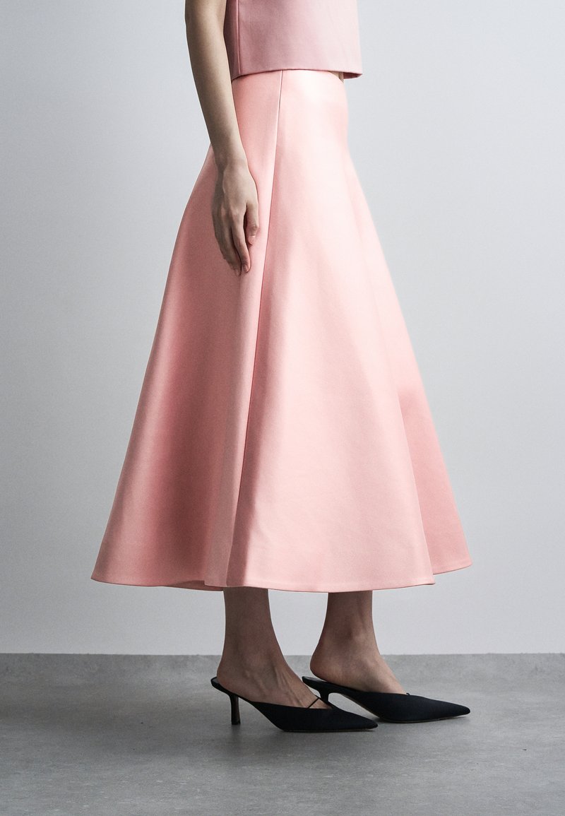 Γυναίκα που φοράει μια ροζ midi φούστα με ψηλή μέση και φαρδιά γραμμή, με το αντίστοιχο τοπ και μαύρα μυτερά kitten heels, στέκεται σε γκρι πάτωμα.