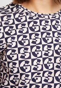 Tričko s krátkým rukávem vyrobené z měkké tkaniny, se námořnickým a krémovým geometrickým vzorem s opakujícími se písmeny 'G' v různých orientacích.