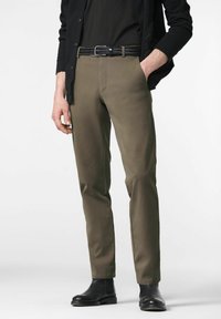 Man in olijfgroene rechte broek met zwarte gevlochten riem, zwart shirt, zwarte cardigan en zwarte leren laarzen, staande voor een witte achtergrond.