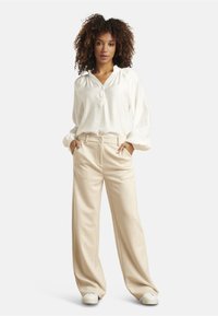 White blouse with a button-up neckline and puffed sleeves; beige wide-leg trousers; model wearing white sneakers; neutral background.