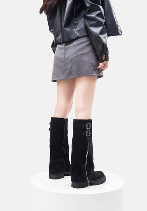 Botas de ante negras hasta la rodilla con detalles de cremallera plateada y dos hebillas; suela rugosa, combinadas con una falda gris y una chaqueta de cuero.