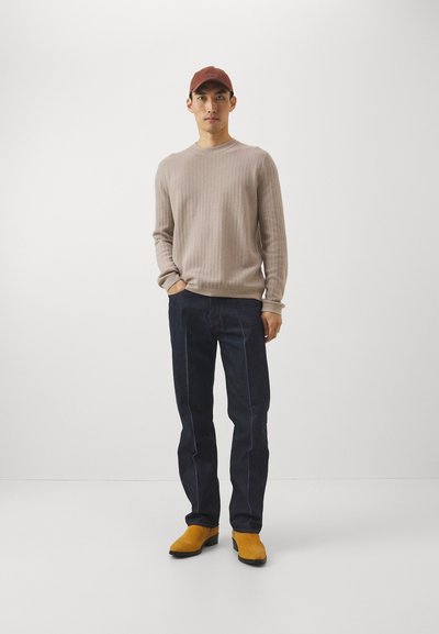 Μπεζ πλεκτό πουλόβερ με υφή, σκούρα τζιν παντελόνια με ανοιχτό μπλε ραφές και κίτρινα σουέτ μποτάκια. Ένα καφέ καπέλο ολοκληρώνει το σύνολο.