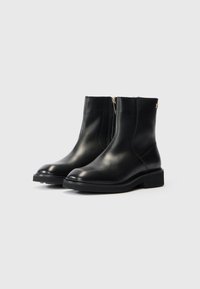 Une paire de bottines en cuir noir avec des zips sur le côté et des talons blocs bas sur un fond gris clair.