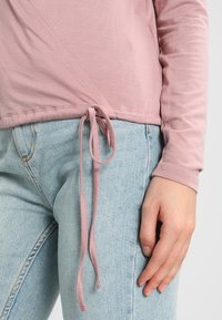 Vaaleanpunainen pitkähiha toppi, jossa on kietaisusuunnittelu ja solmittava yksityiskohta vyötäröllä, yhdistettynä vaaleansinisiin farkkuihin, joissa on hieno tekstuuri.