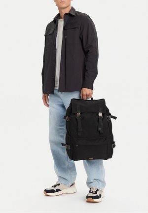 Uomo con camicia nera, t-shirt bianca, jeans azzurri, sneakers nere e bianche, che tiene uno zaino nero grande per la maniglia superiore.