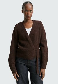 Bruine gebreide wikkelcardigan met een diepe V-hals, textuurstof en een striksluiting in de taille. Gedragen over een witte top en donkere jeans.