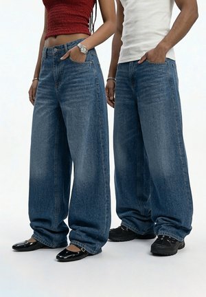 Two people standing side by side wearing wide-legged blue jeans, one in a red crop top and black flats, the other in a white tank and black sneakers.