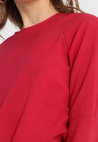 Eine Frau trägt ein simples rotes Langarmoberteil mit rundem Halsausschnitt und weiten Ärmeln.