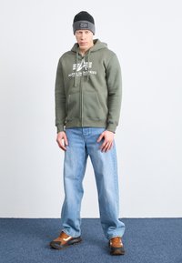Zelená mikina na zips s bielym logom, vyblednuté modré džínsy, hnedo-biele tenisky a čierno-sivá čiapka. Model stojí na svetlom pozadí.
