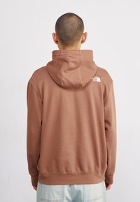 Persona con cabello corto que lleva una sudadera marrón de The North Face y jeans azul claro, de espaldas contra un fondo blanco liso.