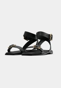 Μαύρα δερμάτινα σανδάλια με φαρδιές λουρίδες, χρυσές μεταλλικές λεπτο μέρειες και ανοιχτή μύτη. Διαθέτουν επίπεδη σόλα και ρυθμιζόμενο τοκά στο αστράγαλο.