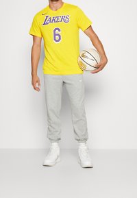Persona con camiseta amarilla de los Lakers #6 y pantalones de chándal grises sosteniendo un balón de baloncesto blanco de Nike frente a un fondo blanco liso.