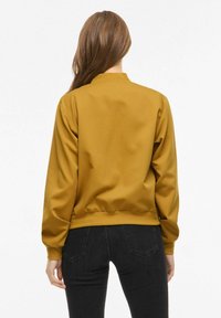 Veste bomber jaune moutarde avec une texture lisse, poignets et ourlet côtelés, et un col simple, assortie à un pantalon noir.