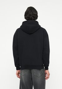 Melnā kapuce ar brīvu piegriezumu, ribotām aprocēm un apakšmalu; izgatavota no mīksta auduma. Aizmugurējā puse nerāda redzamus logo vai grafikas.