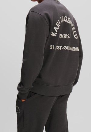 Person iført mørkegrå sweatshirt og bukser med teksten "KARL LAGERFELD PARIS 21 RST-GUILLAUME" på ryggen og sidelommen.
