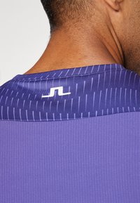Lilla spordipaita, millel on tekstuuriga kangas ja vertikaalsed triibud, ning kontrastne logo kaeluse lähedal.