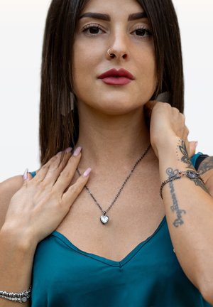 Heart-shaped silver pendant necklace on a dark chain, worn over a teal satin top with a V-neck. Long, straight hair and light nail polish.