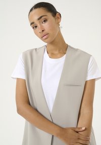 Beige sleeveless vest over a white t-shirt, featuring a narrow chest pocket and smooth texture. Flat, minimalist design with clean lines.