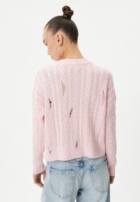 Różowy sweter z warkoczowym splotem, z detalami ażurowymi, z ribbowym wykończeniem i długimi rękawami, zestawiony z jasnoniebieskimi, przetartymi dżinsami.
